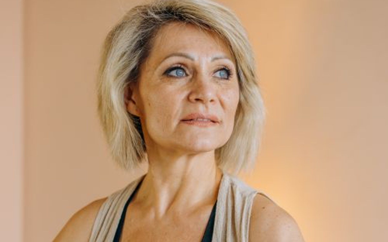 A blonde woman seated on a mat with her hands resting together in a bright room