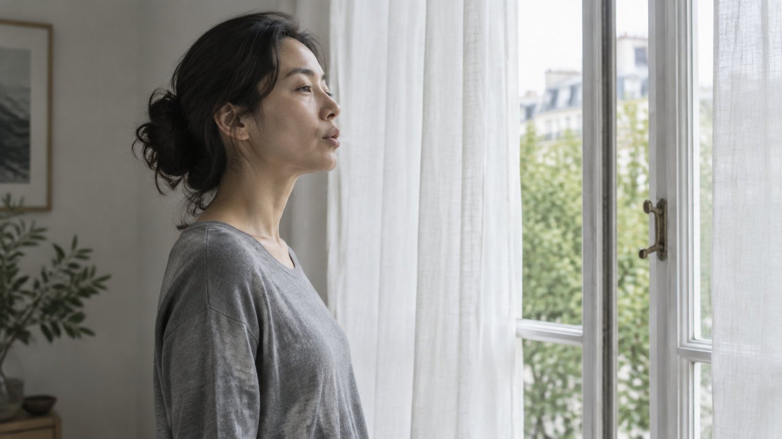 Woman standing by an open window with linen curtains, exhaling slowly through softly pursed lips with relaxed shoulders