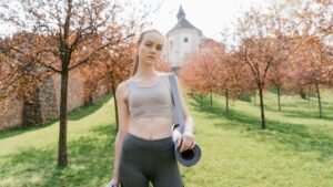 A blonde woman in sportswear carrying a yoga mat in a sunlit park
