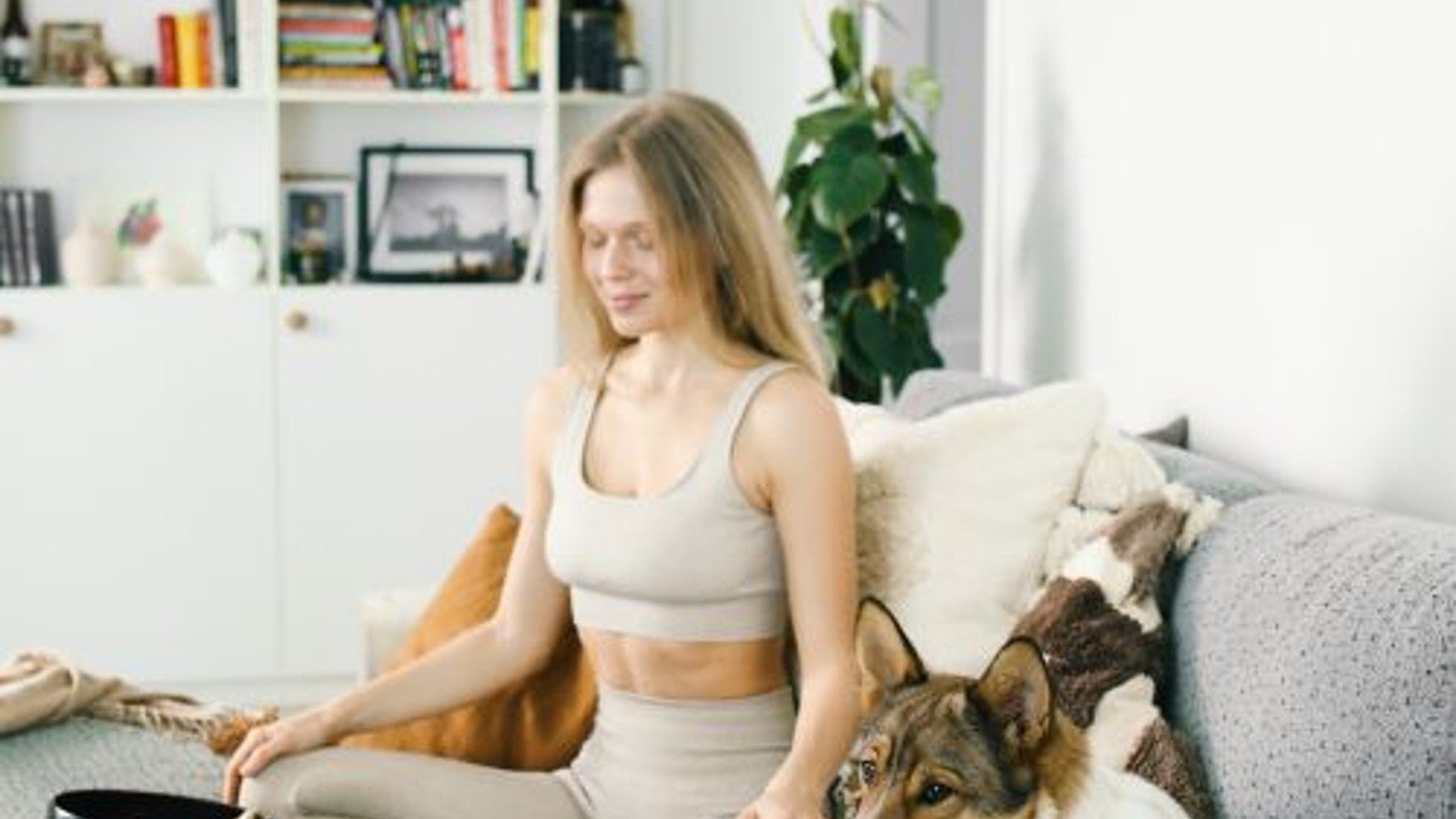 A blonde woman seated on a sofa beside a dog in a bright living room