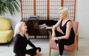 Two blonde women conversing beside a yoga mat, one seated on a chair and one on the floor