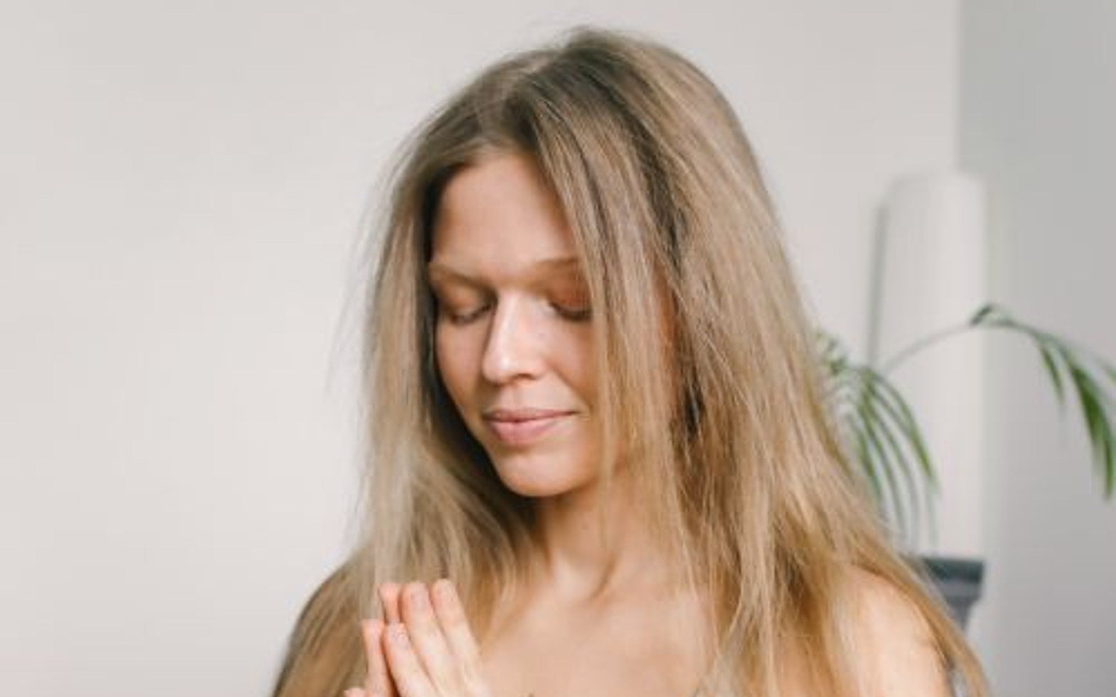 A blonde woman seated indoors with her palms together and eyes closed