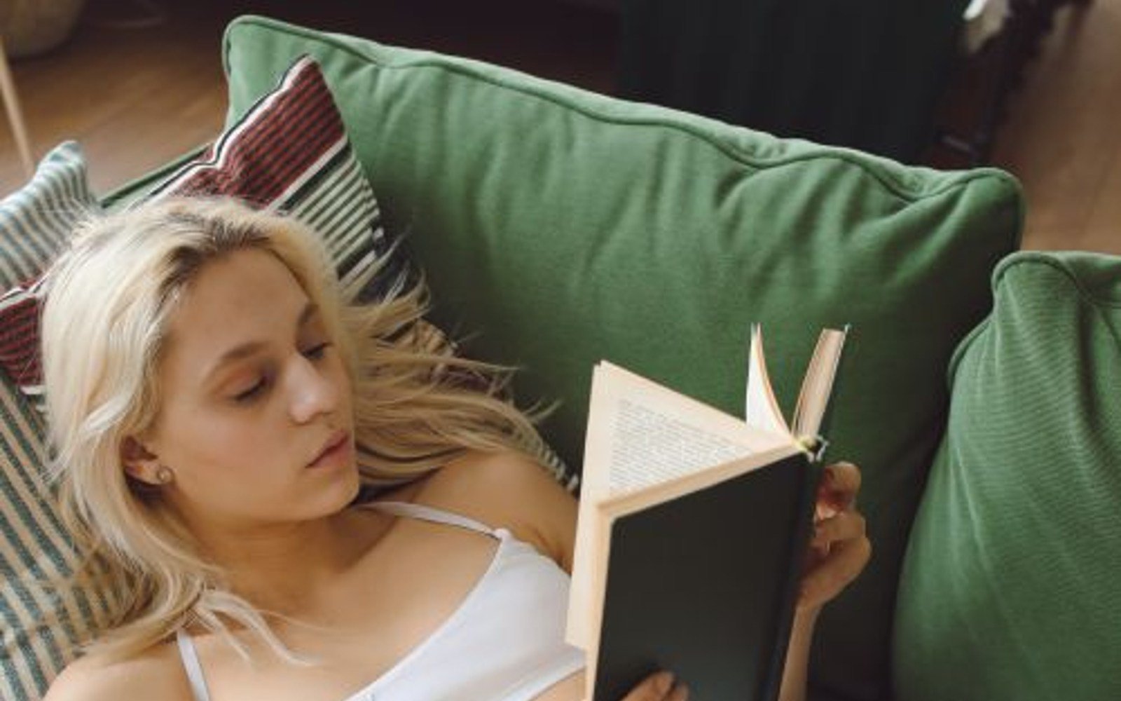 A blonde woman reading a book while reclining on a green sofa