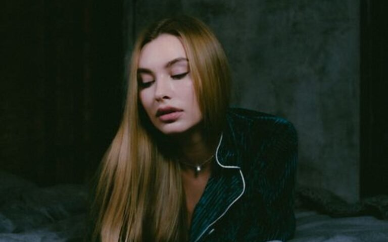 A blonde woman in dark pyjamas resting against pillows on a bed in soft low light