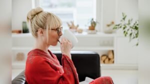 A blonde woman with glasses seated on a sofa holding a white cup in a bright room