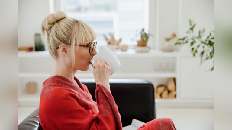 A blonde woman with glasses seated on a sofa holding a white cup in a bright room