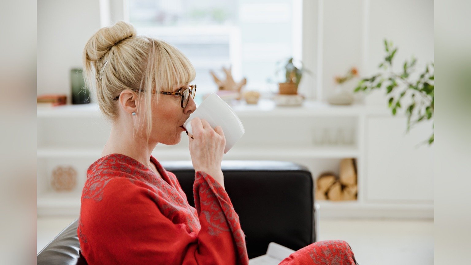 A blonde woman with glasses seated on a sofa holding a white cup in a bright room