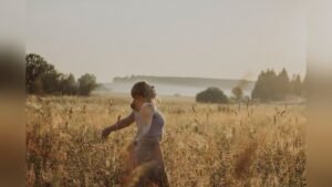 A woman walking through a sunlit grass field with her arms open wide