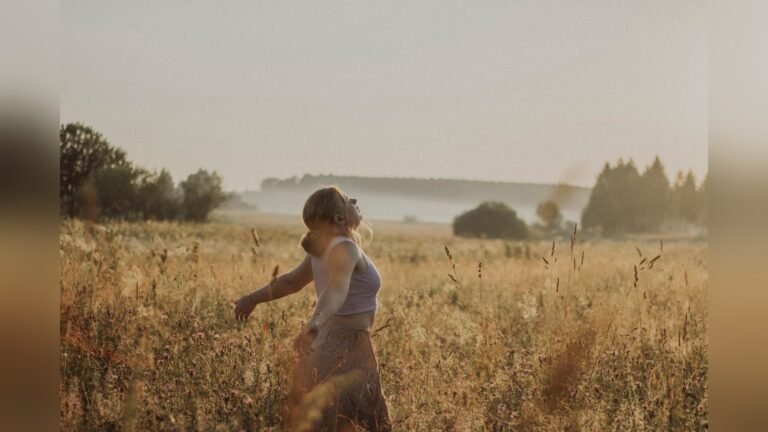 A woman walking through a sunlit grass field with her arms open wide