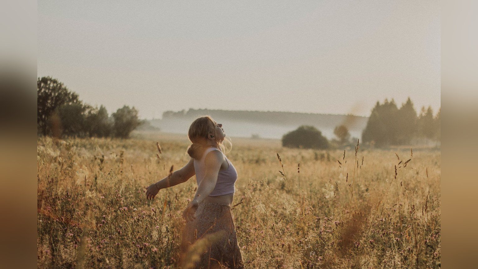 A woman walking through a sunlit grass field with her arms open wide