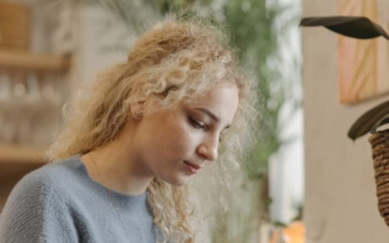A blonde woman seated indoors holding a notebook and pen beside green plants