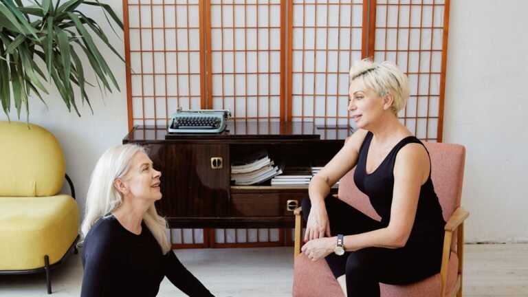 Two women with light hair talking in a wellness room, one seated on a chair and one on the floor