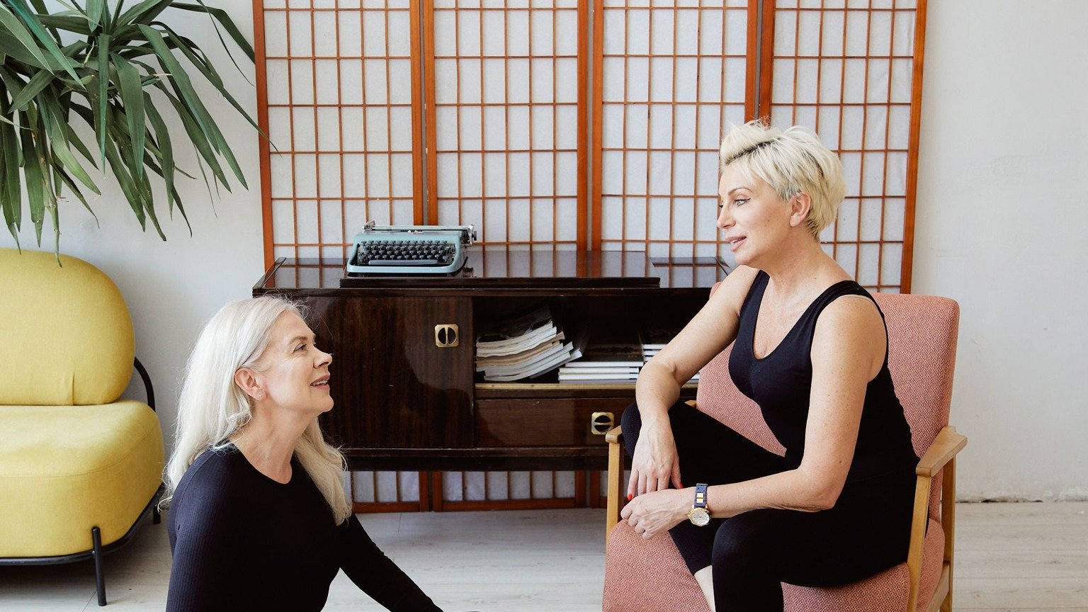 Two women with light hair talking in a wellness room, one seated on a chair and one on the floor