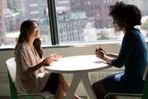 Two women in warm conversation across a table by a large window