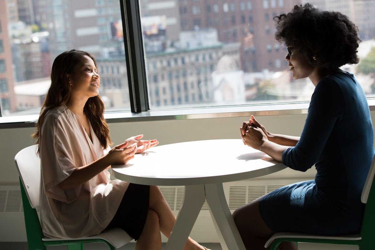 Two women in warm conversation across a table by a large window