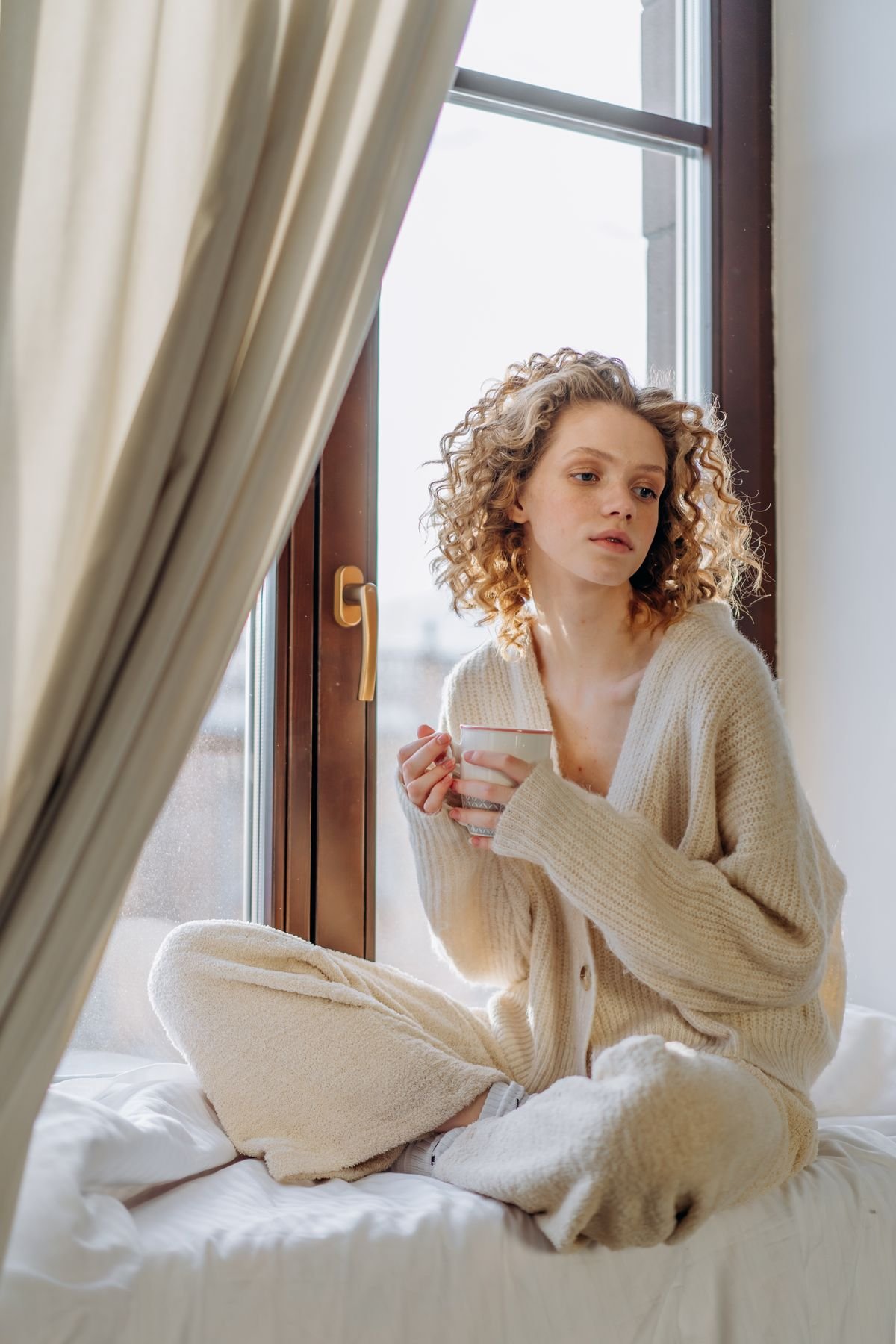 A blonde woman in a cardigan sitting on a window seat in soft light