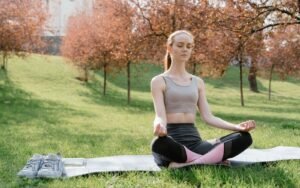 A blonde woman seated on a yoga mat in a grassy park beneath autumn trees