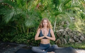 A blonde woman seated on a mat among green plants with her palms together