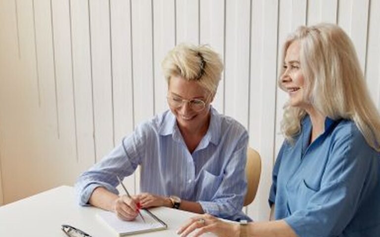 Two blonde women seated at a table with a notebook, one writing and one looking towards her