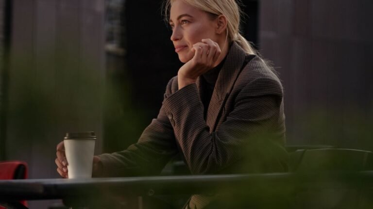 A blonde woman seated at a cafe table looking towards the surroundings while holding a cup