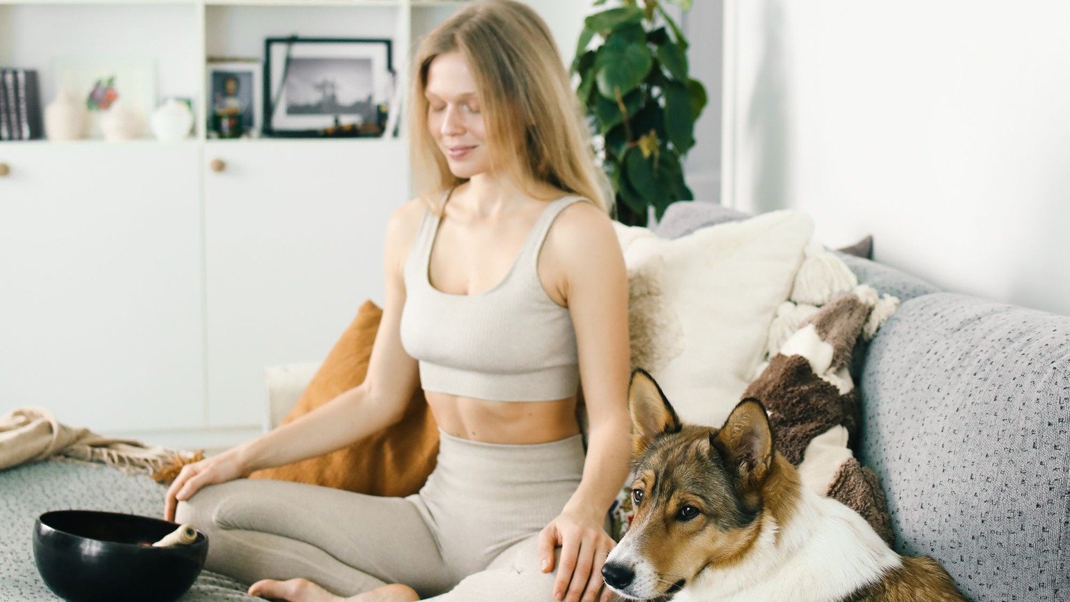 A blonde woman seated on a sofa with her eyes closed, a dog beside her and a bowl nearby