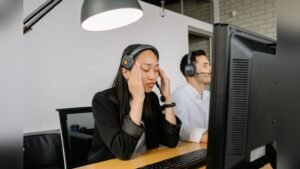 A woman wearing a telephone headset resting her forehead on her hand at an office desk
