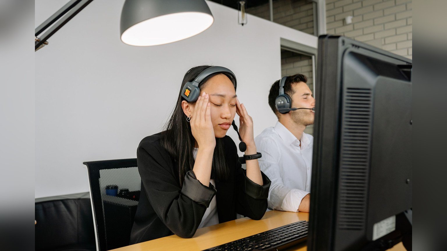 A woman wearing a telephone headset resting her forehead on her hand at an office desk