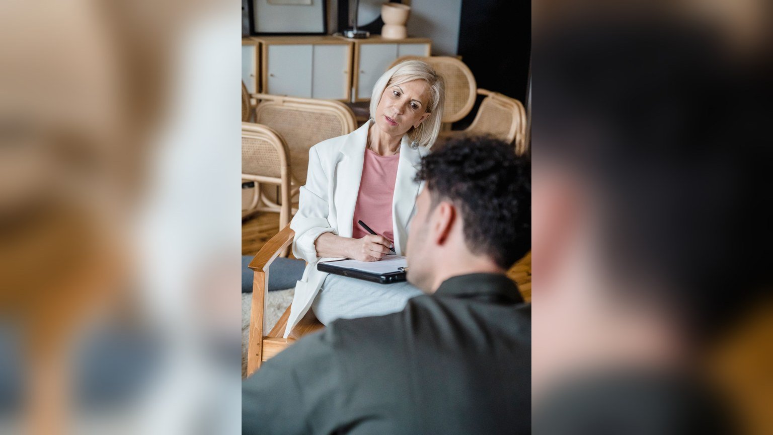 A therapist leaning forward attentively with a clipboard while listening to a client