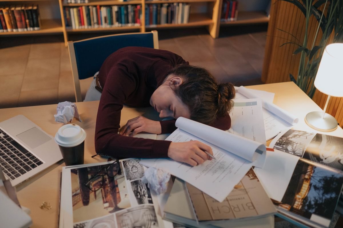 A woman asleep at a desk covered in papers under a warm desk lamp