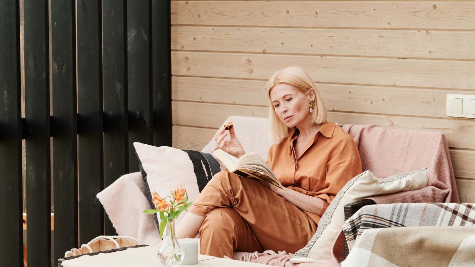 A blonde woman reading on a cushioned sofa on a sunlit wooden patio