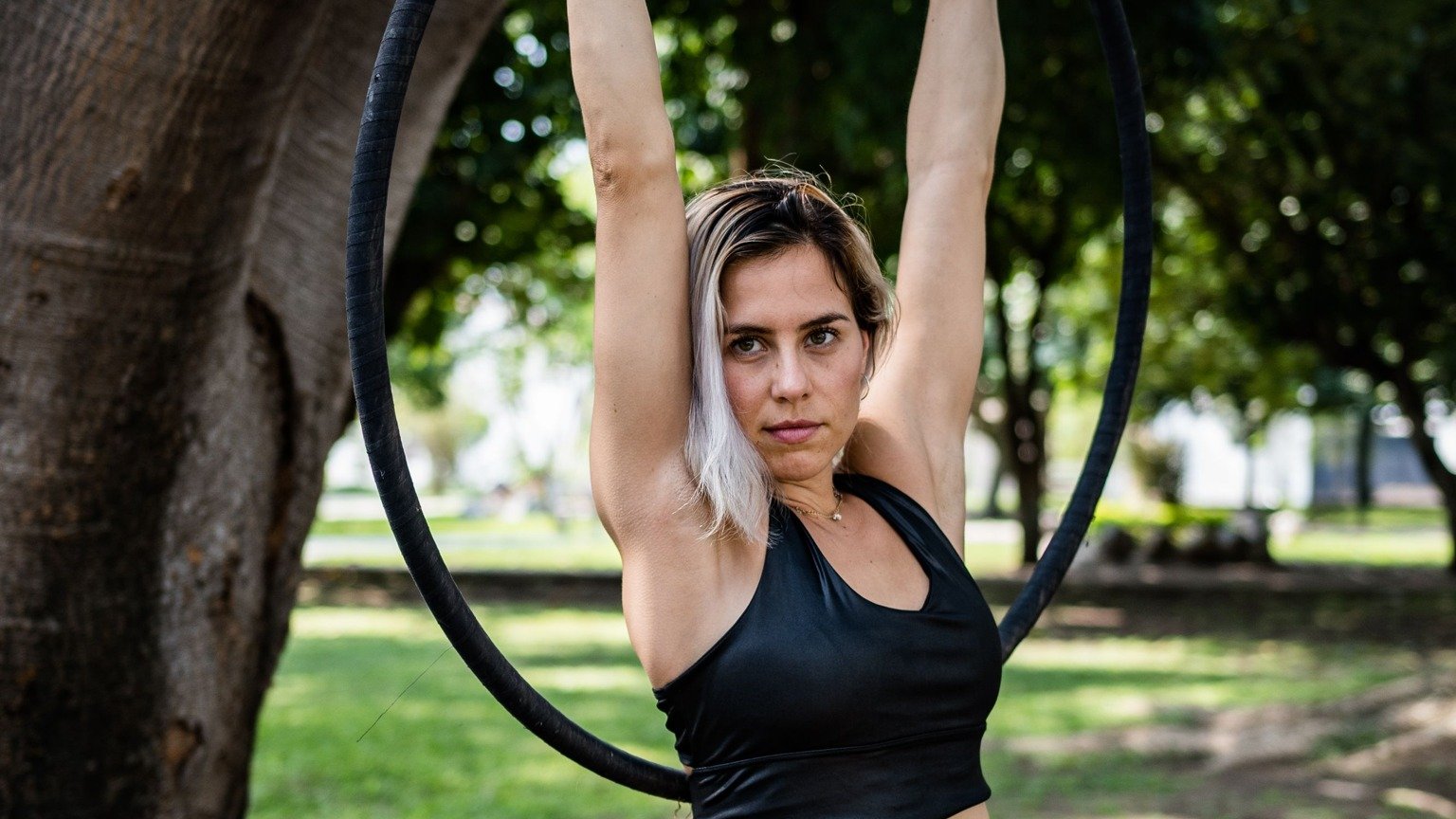 A woman with blonde hair holding an aerial exercise ring above her head in a park